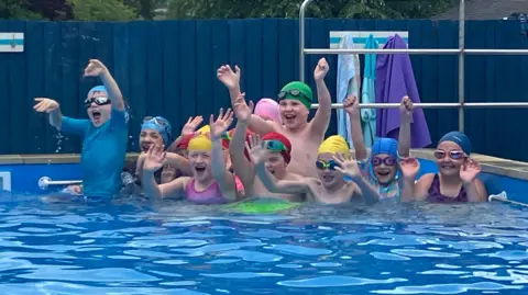 A photo showing a group of children in a swimming pool wearing swimming hats and goggles and they are waving