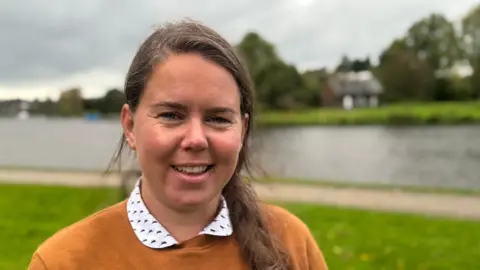 Amy Burns is stood in front of the shoreline in an orange sweater looking directly to the camera