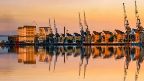 Getty Images Cranes and modern apartment buildings stand at the side of a dock. It is sunset, and the cranes and buildings are reflected in the water. 