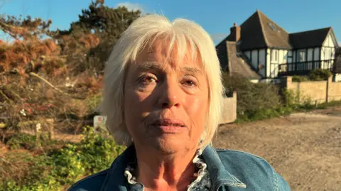 Darren Rozier/BBC A head and shoulders image of a woman wearing a denim jacket while standing on a beach in front of a large black and white house, which is on the right. She is looking into the camera.