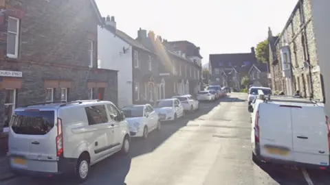 Google Church Street in Keswick is narrow and surrounded by stone and brick terraced houses. There are lines of parked cars on either side of the road. 