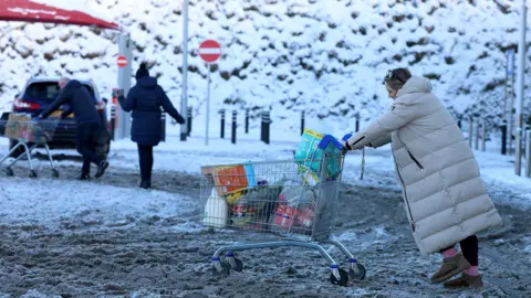 Reuters A woman, dressed in a large, cream winter coat, is pushing a trolley full of shopping in a car park. On the ground, there are large mounds of snow covered in dirt.