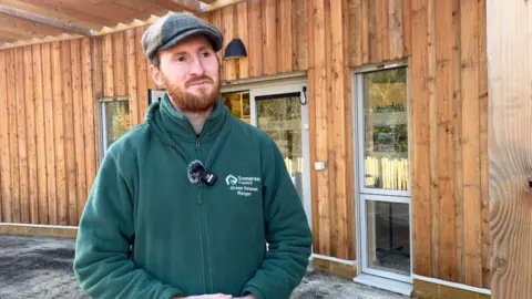 Edd is stood in the middle of the shot, looking to the right of the camera. He's in a green jumper, and is wearing a grey flat cap. Behind him is a wooden-cladded wall.