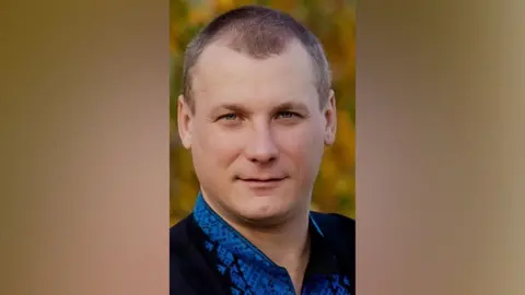 Yevhenii Ivanenko who had short brown hair and is wearing a blue shirt and a black jacket in front of autumnal leaves in a wooded area 