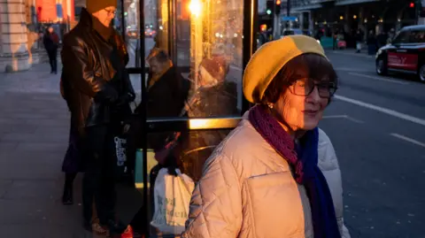 Getty Images An older woman stands at a bus top in the sunset. She has a hat, scarf, glasses and coat on, with people standing behind her.