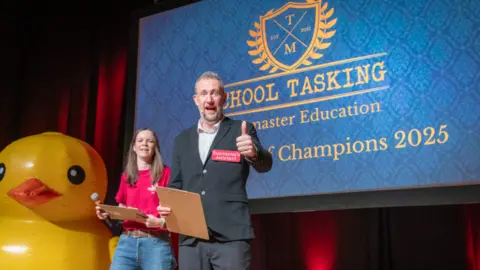 Comedian Alex Horne stands on a stage and looks at the camera doing a thumbs up. Next to him is a woman in a red shirt and blue jeans. On a screen behind them it says School Tasking and Champions 2025, although more text is obscured by Alex Horne. A very large yellow rubber duck is to the left of the picture.