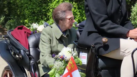Princess Anne sitting in the open carriage holding a bouquet of flowers which has a small Sark flag in it.