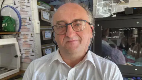 Carl Hawkins wearing a white shirt and glasses stood in the rescue centre with hutches behind him