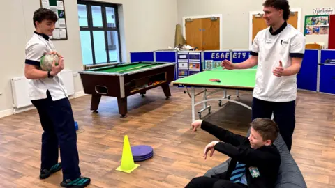Two male OTs and a young pupil are throwing a ball between them in a games room. The OTs are dressed in matching white Ulster University scrubs with a navy blue trim and white name badges. The pupil, who is sitting on a grey sofa, is dressed in a school uniform - a dark coloured blazer, a light blue shirt, and a blue and yellow striped tie. Behind them is a pool table and a ping pong table. 