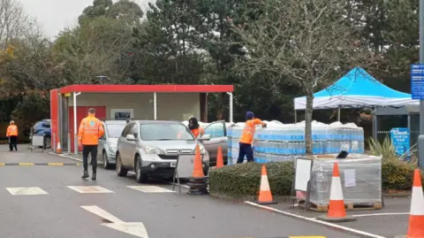 A car stopped next to people in high vis jackets and pallets of bottled water. It is in the car park of tesco in Henley.