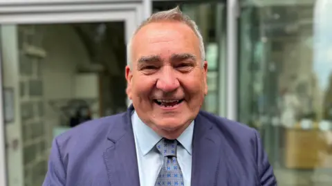 Deputy Simon Vermeulen - A man with grey and black hair, smiling towards the camera. He's wearing a blue suit jacket with a blue shirt and a blue and lighter blue tie.