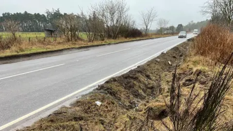 The road verge shows signs of disturbance. There is broken and flattened ground. On the road are a car and van. The sky is grey.