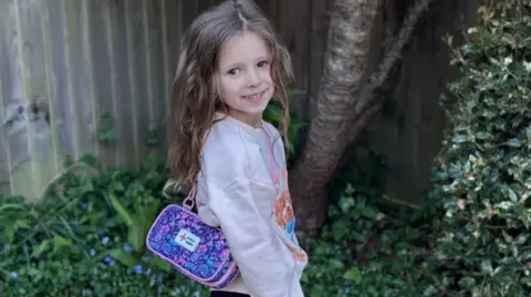 Submitted Margo standing in the garden, with her epipen case over her shoulder, which is purple and pink in colour. There are bushes around her and she has on a beige jumper and dark trousers