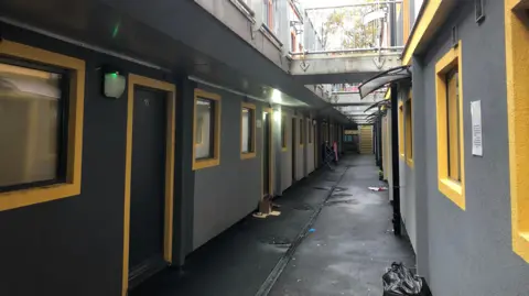A passageway between two rows of two-storey flats with yellow rimmed windows and grey walls. There is a black plastic rubbish bag in the foreground and yellow steps at the end of the passageway. There are connecting walkways on the first floors between the two rows.