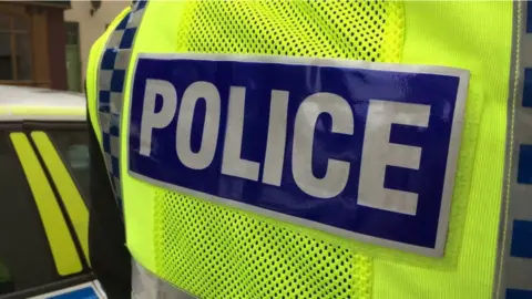 The back view of a yellow British police vest with the words police in white block lettering on a blue horizontal strip.