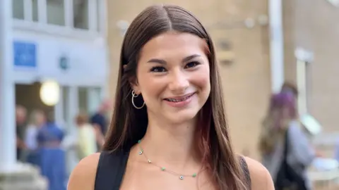 A young girl with long brown hair wearing hoop earrings and braces is smiling at the camera.