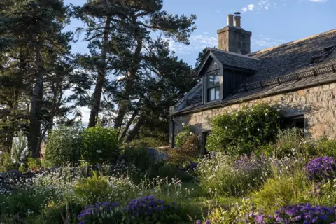 Ben Addy Ardoch rises from a lush green garden, a stone-built cottage surrounded by wildflowers and towering trees