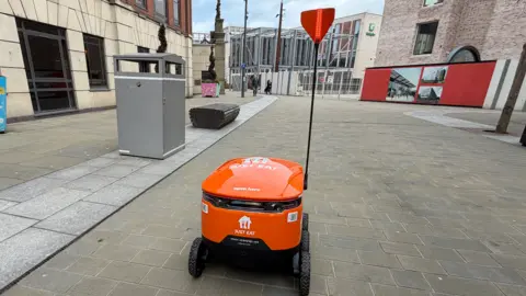 BBC/Jim Scott The orange coloured robot, with Just Eat branding in white, is travelling towards the city's Keel Square. It has six chunky rubber wheels and a large pole which carries a flashing orange flag to indicate it is near. In the distance is the under-construction Culture House. 
