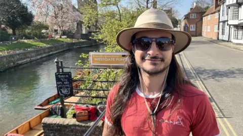 Phil Harrison/BBC A man in a red t-shirt, sunglasses and a wide-brimmed hat smiling at the camera. A river with some wooden punt boats at the side of the water is seen behind him. It is a sunny day.