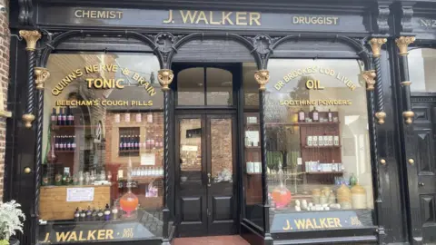The outside of John Walker's shop in Stockton. Signs in the left window read 'Quinn's nerve and brain tonic' and ' Beecham's cough pills'. On the right gold letters say 'Dr Brookes' cod liver oil' and 'soothing powders'.