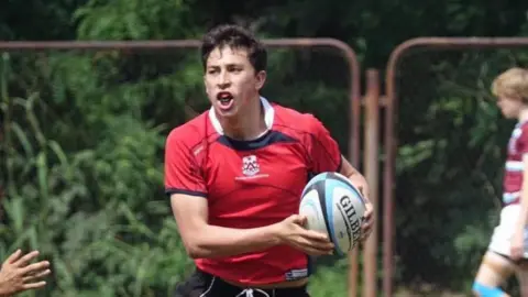 Jelenyela Pena Seb Pena wearing a red jersey has a rugby ball in his hand. He appears to be in motion, and there is a rusty fence behind him.