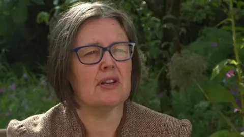 Alison Norris with chin-length brown hair in blue glasses wearing a brown blazer while sitting in a garden surrounded by greenery