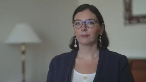 A woman with medium length dark hair, tucked behind her ears, is sitting in a room facing a window, with the light reflecting from her round, dark glasses. She is wearing a white blouse and navy jacket, with a chain around her neck and dangling earrings. She is in a room with a lamp and a sofa visible behind her.
