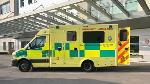 Getty Images A yellow ambulance with green patches. The words "Emergency ambulance" are visible in red lettering at the top of the side of the ambulance. The trust's name is in black writing half way down the side. A rear door, with red stripes, is open. The ambulance is parked outside a hospital.
