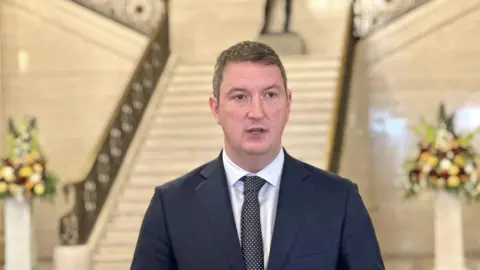 PA Finucane with short dark hair, he is wearing a navy suit, spotty tie and a white shirt.