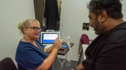 City of Wolverhampton Council A female nurse, wearing glasses and a blue uniform, is sitting at a desk with medical equipment set out. She is looking at a man with a beard who is wearing a dark-coloured top. He is sitting opposite the nurse.