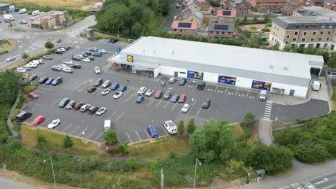 An aerial photograph shows a Lidl supermarket and its surrounding car park on the outskirts of Beverley. Dozens of cars are parked in marked bays around the store, which sits beside a busy roundabout and main road. Residential housing, trees and nearby retail units can be seen around the site, while footpaths run along the edge of the car park.