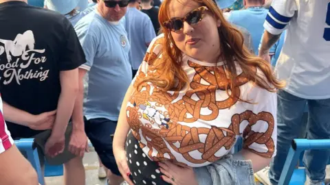 Helen Powell is smiling as she stands amidst Manchester City fans. She is visibly pregnant and has her hands on her bump. She has long red hair and is wearing a white top with a pretzel pattern.