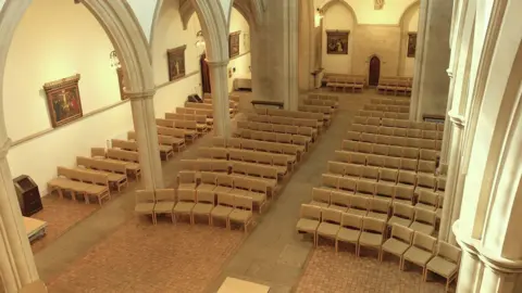 The interior of a church. It's made of light brown stone with large archways on either side, and rows of beige chairs in the middle