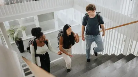 Three students wearing rucksacks on their backs chatting happily with each other as they climb steps inside a building.