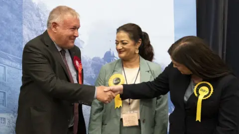 Davy Russel wearing a suit and red rosette shakes the hand of an SNP candidate in a yellow rosette