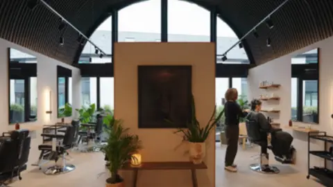 Lewis and Hickey An artist's impression of a hair salon which makes use of the space within a railway arch. The frontage and roof of the room is curved with a series of chairs and mirrors down each side along with a table with potted plants on it.