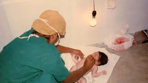 SOUTH TEES HOSPITALS NHS TRUST A woman in medical scrubs is leaning over a new-born baby on a white mat. A naked lightbulb can be seen above them.