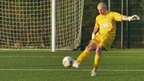 The HALO Trust Kieran McCulloch in goalkeeper strip, kicking a ball on a football field with a goal in the background.