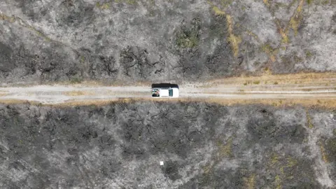 A white pick-up truck drives along a gravel track surrounded by blackened heath following the severe fire 12 months ago