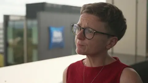 Bev Craig has short brown hair and is wearing dark framed glasses and red top.