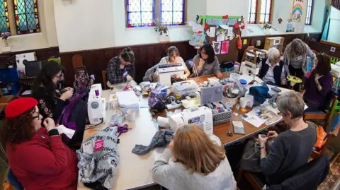 Farnham Repair Café Ten people sat around a table using sewing machines to repair clothes. They are in a church hall with dark wood panels half way up the walls. There are stained glass windows in the wall