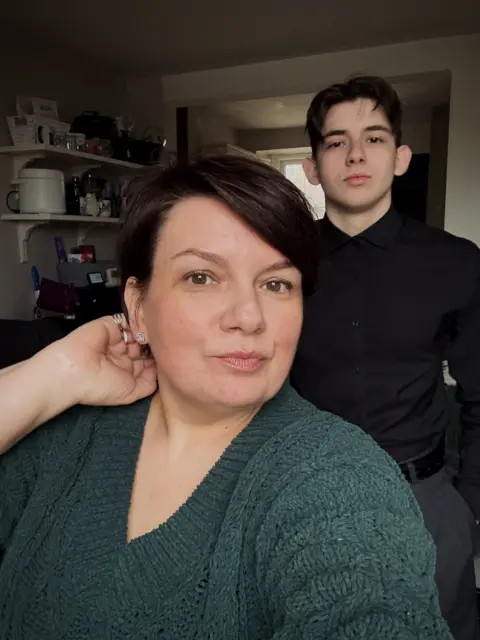 Oles Kuzomko On the left Liudmyla, is looking at the camera wearing a dark green knitted jumper. Her hair is short and dark. Oles is standing on the right wearing a black shirt and trousers. His hair is short and dark brown. 