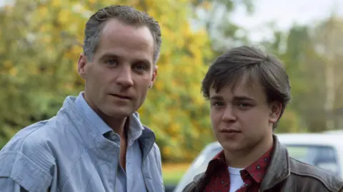 Lord Michael Cashman (left) as Colin Russell and Gary Hailes (right) as Barry Clark in EastEnders. The man on the left has brown/grey hair and is wearing a blue shirt. The man on the right has brown hair with a red shirt and brown leather jacket.