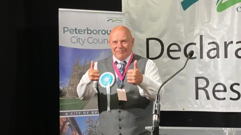 Shariqua Ahmed/BBC John Robert Bolton wearing a white shirt and grey waistcoat, giving a thumbs up to the media.