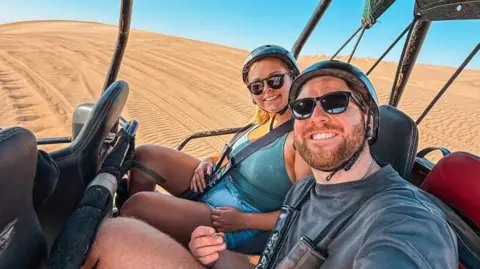 That Travel Couple A selfie of Hannah Bird and Charlie Camper strapped into a dune buggy on a vast plain of desert. They are both wearing black helmets and sunglasses, and smiling at the camera.