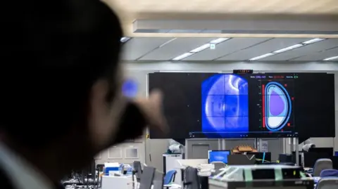 Man pointing across a room full of computers at a large screen which is showing images from the inside of fusion reactor.