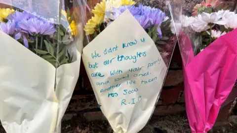 Martin Barber/BBC Three bouquets of flowers which have been placed against a brick wall. One bouquet has bright pink wrapping and the other two (in the centre and on the left) are wrapped in a cream colour paper. Written on the bouquet in the centre a message reads: "We didn't know you but our thoughts are with your family and loved ones. Another life taken too soon. RIP."