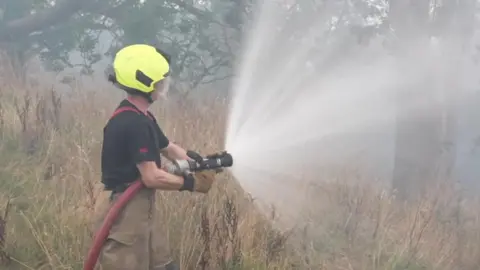 BBC A male firefighter wearing a yellow helmet, a black T shirt and beige trousers holds a large red hose and sprays water at moorland grass