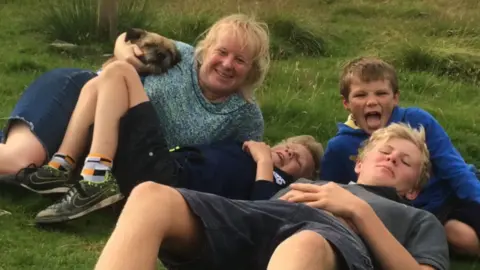Harris Trust A boy with blonde hair, lying on the ground, leans back against a dark-haired boy with another blonde haired boy lying next to them and a woman with blonde hair cradles a small brown dog. All are smiling at the camera
