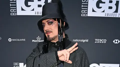 Boy George is wearing a pinstripe jacket and a black bucket-style hat with tassles. He has facial hair, colourful eye make-up gems around his eyes. Behind him is a black wall with the logo 'British LGBT Awards' on it. He is making a peace sign with two fingers.